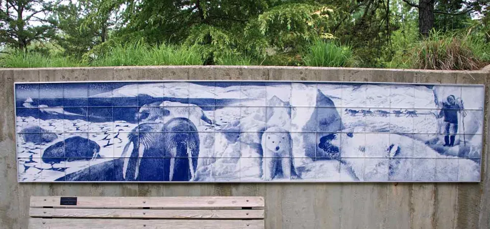A wide, light wooden bench with a black metal frame sits in front of a long, rectangular mural on a concrete wall. The mural, rendered in shades of blue and white, depicts Arctic wildlife, including walruses and polar bears, and a human figure in winter gear. Green trees and foliage are visible above the wall in the background.