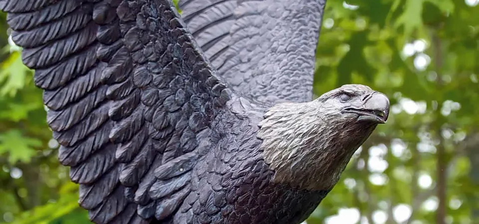 A bronze statue of a Bald Eagle mid-flight, its wings raised and flapping in the wind. A forest of dense trees is visible in the blurred background.