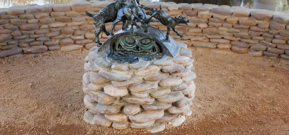 A bronze sculpture of three goats, with swirling forms and intricate details, atop a circular pedestal of stacked, light-colored stones. In the background, a calm body of water and two concrete pillars of a bridge.