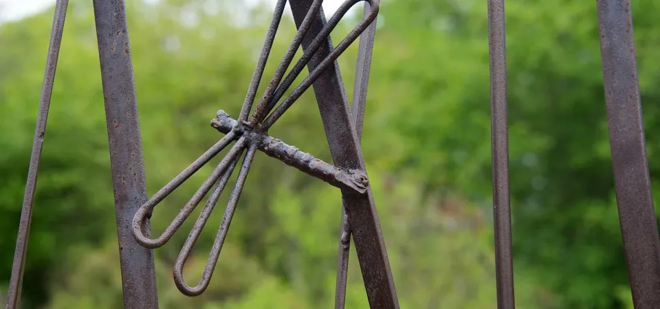 A closeup on a part of a metal art installation that showcases a small, metal dragonfly appearing mid flight in front of some tall cattail stalks. There is a green forest visible in the blurred background.