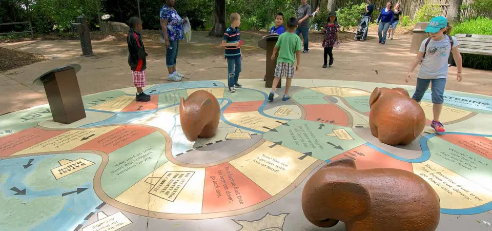 A vibrant outdoor board game with colorful pathways and large brown game pieces shaped like Elephants on a paved circular area. Several children and adults are gathered around and on the game, with lush green trees and foliage in the background, suggesting a park or zoo setting.
