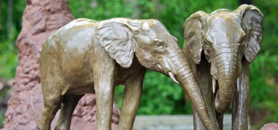 A small bronze statue of two Elephants standing together, one reaching its trunk out towards the other. Mounds of mud and a termite mound are placed around them.
