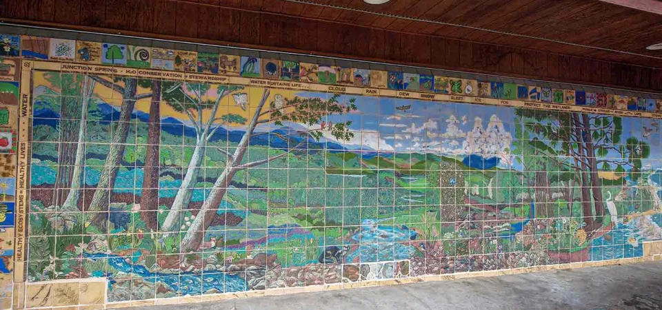 A vibrant, large-scale mural made of square tiles depicts a lush, expansive landscape. On the left, tall trees with light-colored trunks and green foliage frame a scene that transitions into a wide-open field in the center. A light blue river flows from the left foreground to the center. The mural extends across a long wall under a brown overhang.
