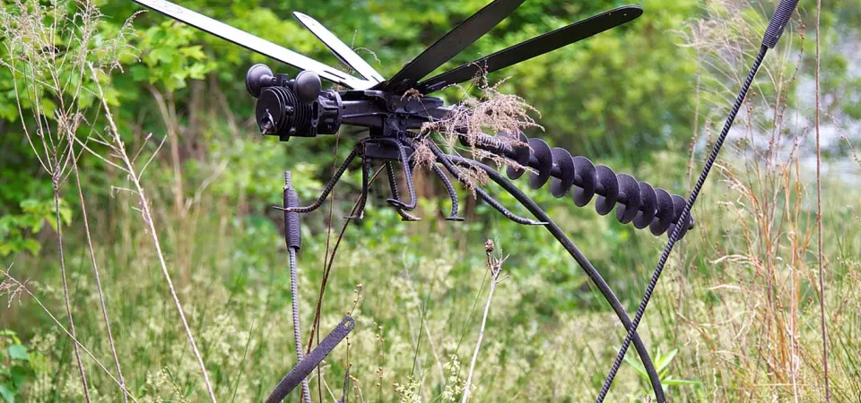 A large Dragonfly sculpture created from recycled metal pieces. It is attached to a metal bar that makes it look like it is mid flight amongst the tall grasses of a swamp.