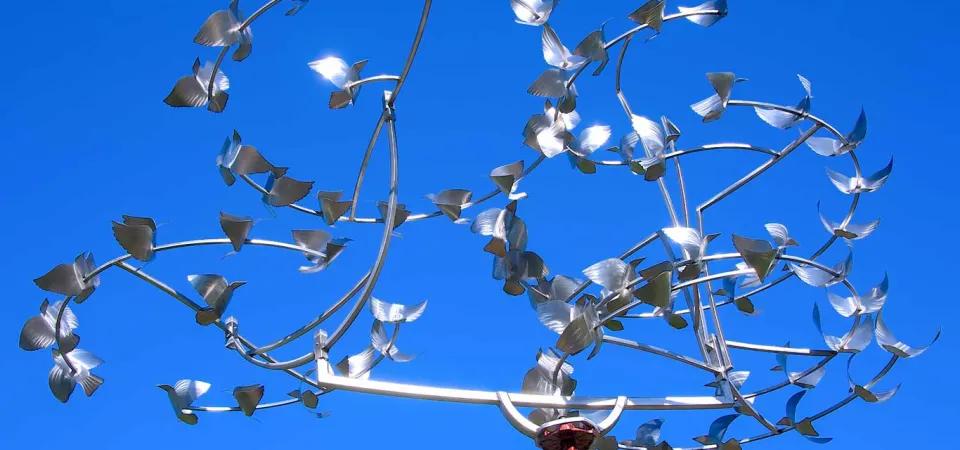 A low-angle shot captures an art installation that is designed to spin in the wind like a weather vane against a clear, vibrant blue sky. It is made of several spiraling, circular, silver tubes that are lined with smaller, silver shapes that look like wings. As the piece spins, it looks like butterflies fluttering in a circle. The central support structure is a reddish-brown pole, visible at the bottom of the sculpture. 