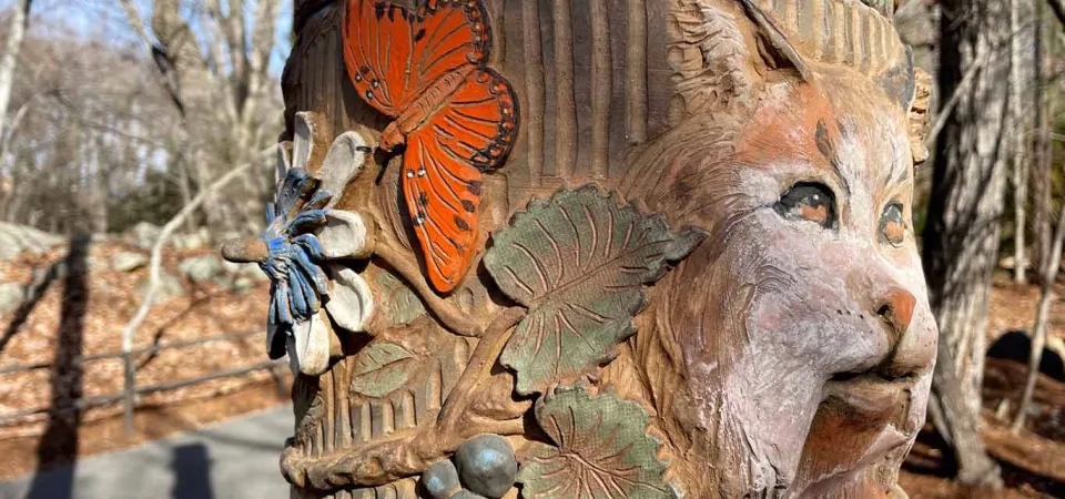 Zoomed in view of a tall, round, wooden pole that is covered with different colorful images that have been carved into the wood. This image features carvings of a lynx, butterflies, and various plants and flowers.