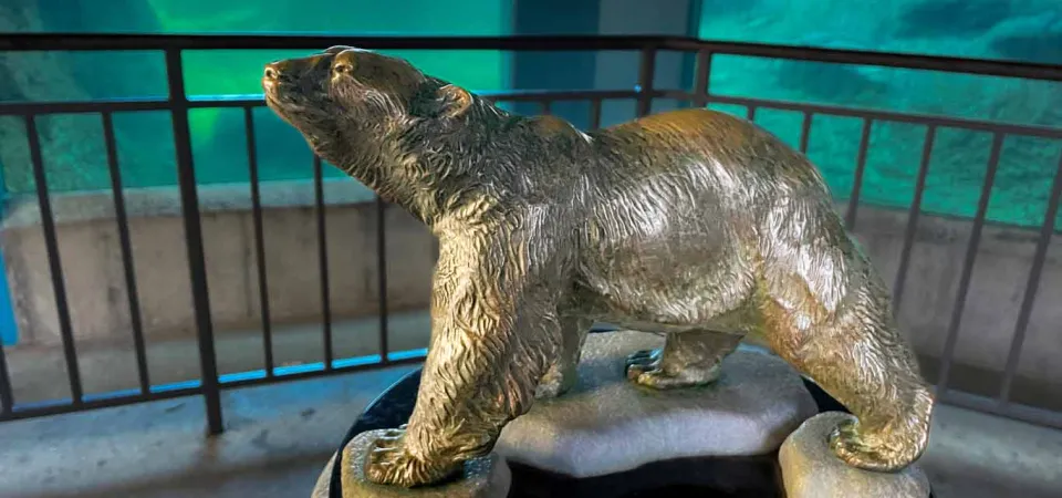 A small bronze statue of a Polar Bear walking on ice that has been placed on top of a pillar. There is a metal handrail lining a ramp that leads to a glass viewing area of an animal habitat pool in the background.