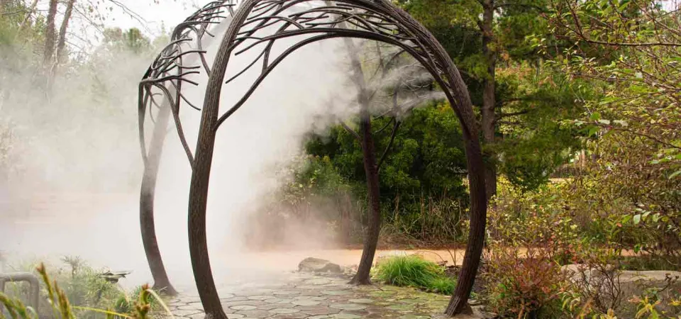 An intricate, domed archway with a tree branch design, that spans a sidewalk that is lined with a variety of plants. There is water being sprayed down in a fine mist to keep those who walk underneath it cool.