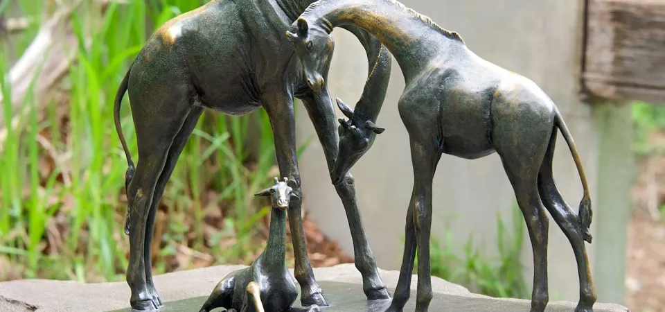 A small bronze statue of two Giraffes standing in front of each other, their necks bending down and crossing each other to look at a baby Giraffe that is laying between them. This artwork is affixed to the top of a large stone.