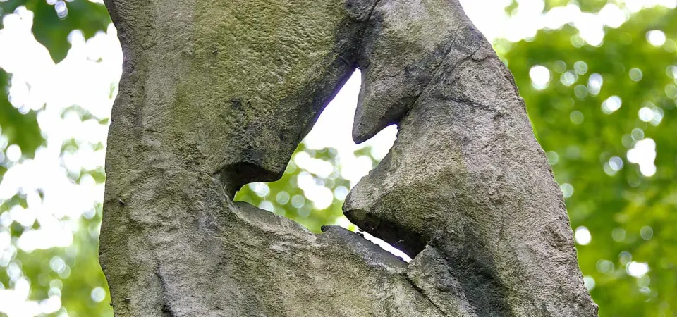 A large stone with the shape of a bird that appears to be in flight carved into the middle of it. A canopy of trees and a clear sky are visible through and around the stone.