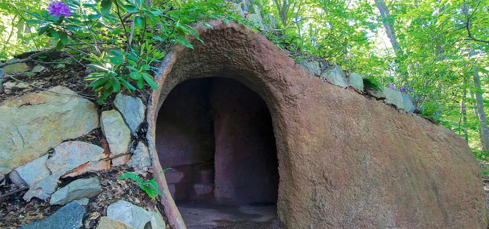 A whimsical, hobbit-hole-like earthen structure with a rounded archway entrance is nestled into a hillside in a lush green forest, blending seamlessly with the surroundings. To the left of the entrance, a stone wall is integrated into the structure, and a vibrant purple rhododendron bush with green leaves blossoms atop the earthen roof.