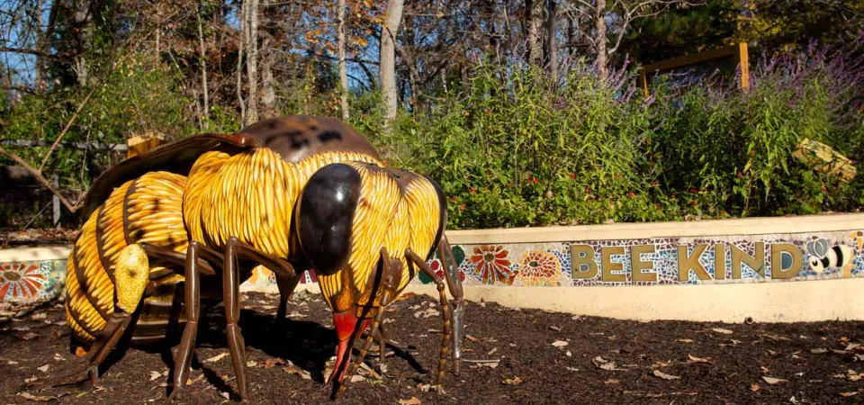 A gigantic statue of a worker bee stands on the ground at the Garden Friends Playground with its wings tucked back. A mosaic wall stands behind it with tiled words that read "Bee Kind" and various small flowers.
