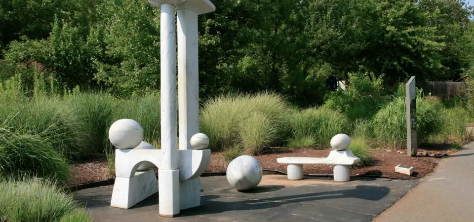 Zoological egg rest art installation which is comprised of minimalist white stone sculptures in a lush green outdoor setting, with a tall central piece, spherical elements, and a bench-like form beside a paved walkway.