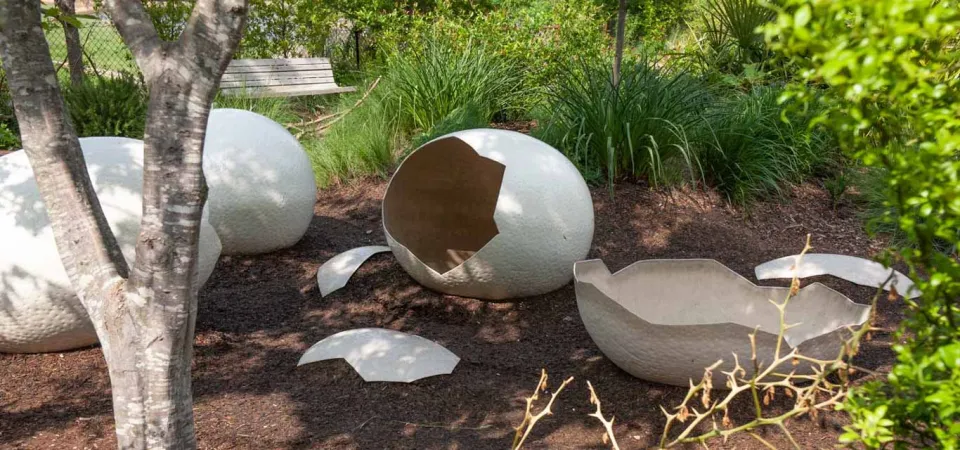 Three oversized, cracked white eggshells are scattered on a bed of dark mulch or dirt in a garden setting, suggesting a recent "hatching." The shells have a textured surface like rough concrete or stone. The scene is surrounded by lush green foliage and tall grass, with the base of a small tree in the foreground to the left and a wooden bench visible in the background. This is likely an outdoor art installation or a zoo/garden exhibit.