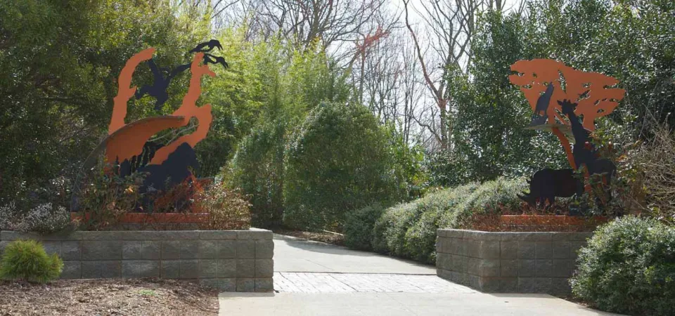 A panoramic view of the entrance to an outdoor path flanked by two large, metal sculptures of animals in dark brown or black against a rusted orange background, depicting scenes like a giraffe eating from a tree on the right, and other African animals, possibly including a lion, on the left. The path is brick and concrete and leads between two low stone or concrete planter walls topped with shrubbery.