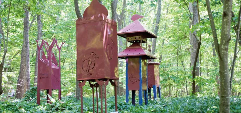 A group of tall, artistic metal sculptures resembling red-brown lanterns or towers, featuring animal cut-outs, are displayed outdoors in a dense wooded area.
