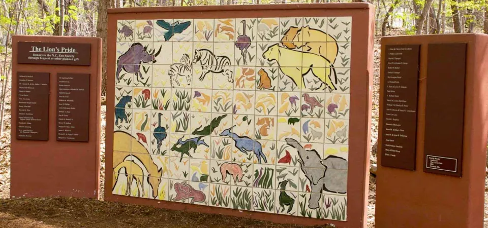  A colorful outdoor mural made of a large grid of painted ceramic tiles depicting various African animals, including a rhino, lion, elephants, zebra, and several birds, set against a background of plants. Flanking the tile mural are two brown panels with white text; the left panel is titled "The Lion's Pride."