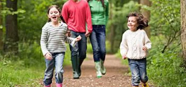 A group of four people, possibly a family, consisting of a man, a woman, and two small girls, walks along a dirt path in a wooded area. The girls are running ahead of the adults and appear to be laughing. The adults are walking behind them and may be smiling. The individuals are wearing casual clothing, including jeans and sweaters.