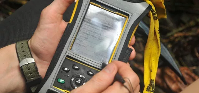 View of someone's hands holding a SMART spacial monitoring and reporting tool, which is a small, black handheld device the size of a cell phone.