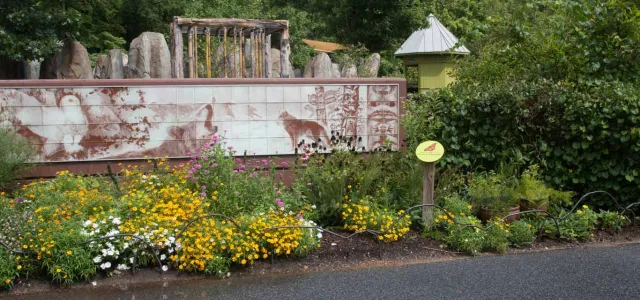 A low-angle shot captures a section of a zoo exhibit. In the foreground, a bed of yellow and white flowers borders a dark asphalt path. Behind them is a long, horizontal mural depicts what appears to be various animals in a rustic, almost weathered style. Above and behind the sign, a structure resembling ancient ruins with multiple stone columns is visible. The background is filled with a thick canopy of green trees.