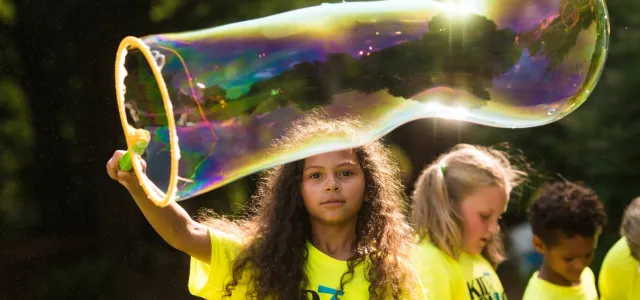 A young girl with long curly hair uses a wand to blow a giant bubble. There are trees and a group of other kids out of focus in the background.