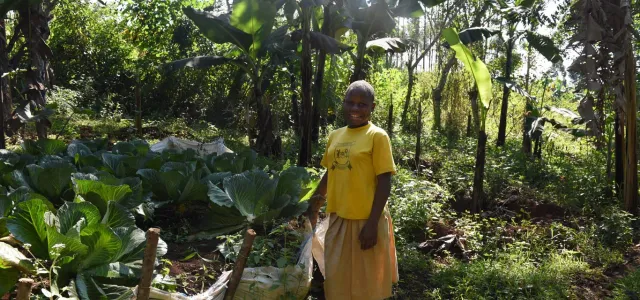 A person with dark skin and a shaved head stands smiling in a sunny, lush green garden. They are wearing a bright yellow t-shirt and a tan skirt. To their left, large, leafy green plants are growing in rows in some light-colored sacks that have been repurposed for gardening. Tall trees and various green plants fill the background, suggesting a tropical environment. 