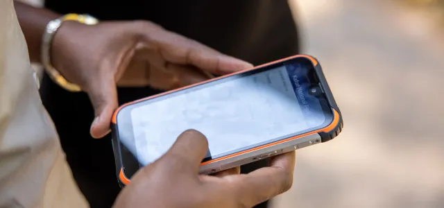 A pair of hands holding a device that looks like a cellphone. Another person, visible from the waist down, wearing black pants, is standing next to them.