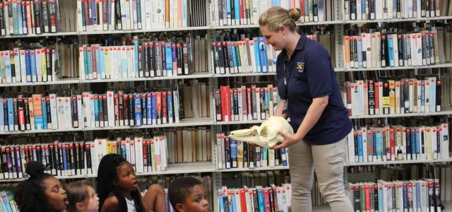 A woman with a blonde bun and navy NC Zoo uniform shirt stands in front of a group of children holding a large skull. A wall of bookshelves filled with books fills the wall beside her, giving the impression of an educational presentation at a school library.