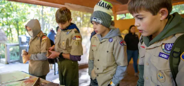Four young people appear to be gathered around a table outdoors. They are wearing tan boy scout uniforms that are covered with patches and appear to be listening to another individual who is not visible to the viewer. On the table are various objects, including what appear to be drawings and graphs. The setting looks like a covered outdoor area with stone pillars or walls in the background.