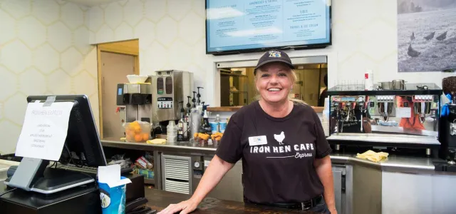 A woman wearing an Iron Hen Cafe hat and uniform leans casually on a wooden restaurant counter next to a cash register. There are food items, kitchen machinery, and kitchen visible behind her. A menu hangs on the wall behind her head.