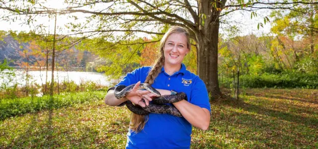 A woman with a long, blonde braid, wearing a blue collared shirt is standing next to a lake in the woods. She is smiling at the camera and holding a very long, black snake in both hands as it wraps around her hands and forearms.