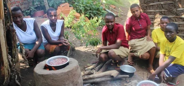 Six young women or girls are crouching around a traditional outdoor cooking fire set up on the ground, likely in a rural African community. In the center, a cooking pot filled with red beans sits on a raised clay or stone stove. The girls are looking towards the viewer, wearing a mix of school uniforms (white vests and skirts/wraps, or maroon shirts) and yellow t-shirts. More cooking pots and wood are visible on the ground.