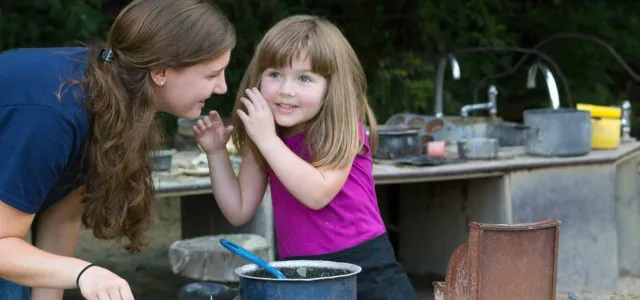 An Educator is talking with a young white girl in pink at the Mud Cafe with a steel counter behind her that is littered with kitchen utensils.