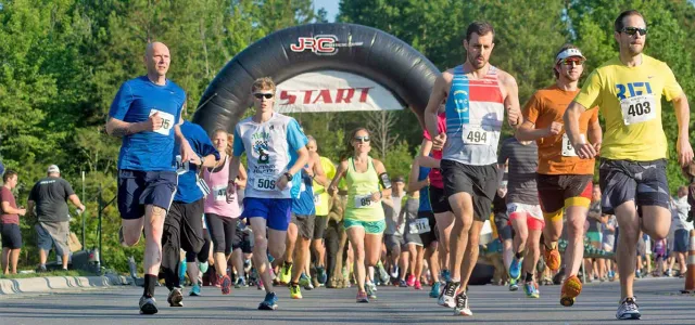 A large group of runners is starting a road race, moving forward beneath a large black inflatable arch labeled "START". The runners are wearing various brightly colored athletic gear.