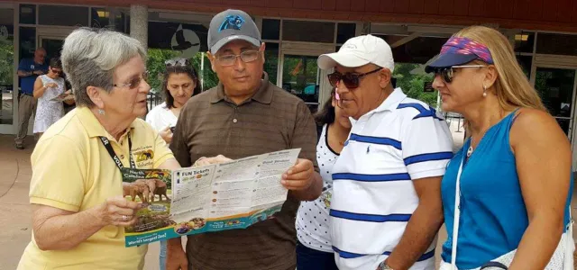 A zoo or park employee in a yellow shirt is helping three visitors by pointing to information on a folded map or brochure. They are standing outside near the entrance to a building.