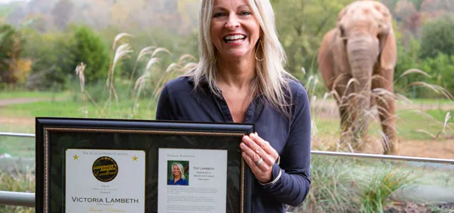 A smiling woman with long blonde hair, wearing a dark blue shirt, stands outdoors holding a framed document or award with two sections. In the background, behind a low metal railing, a young, reddish-brown elephant is visible standing in a grassy enclosure.
