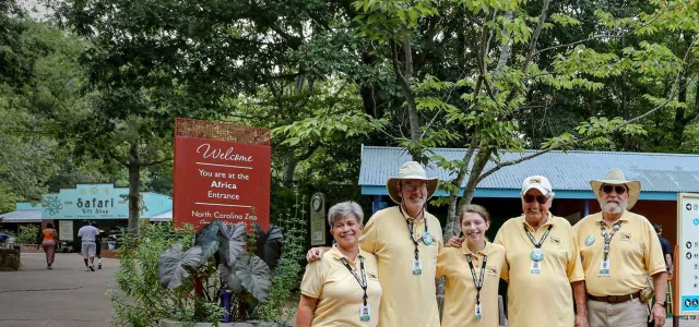 Five Zoo volunteers wearing yellow shirts and black lanyards posing, arms around each other, for the camera in the lush Africa Plaza.