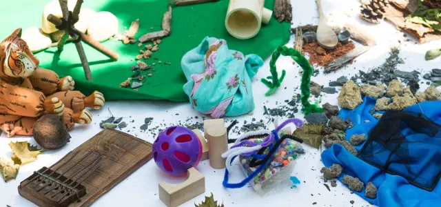 A table covered in a white cloth and littered with a variety of craft items including a toy tiger, green clay, fuzzy pipecleaners, pinecones, green paper, sand, sticks, wooden blocks, and a blue plastic material.