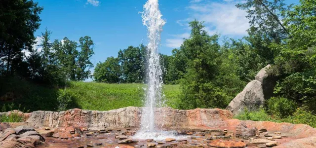 A fountain or geyser feature spraying a tall column of water high into the air against a bright blue sky with scattered white clouds. The water erupts from a base of reddish-brown and tan rocks, which are wet and surrounded by smaller, dark stones. The background is a vibrant green landscape with lush trees and a grassy hillside under the sunny sky.