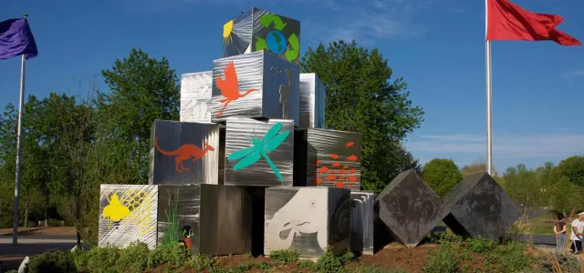 A striking silver cube sculpture, representing themes of conservation and nature, stands on a grassy mound near a pathway. The cubes are adorned with brightly colored environmental and wildlife graphics.
