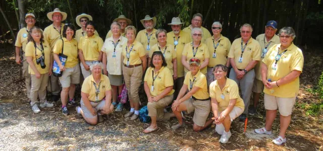 A group of approximately twenty adults, all wearing yellow polo shirts, stands together posing on a gravel parking lot with a dense forest in the background.