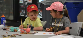 Two children sitting at a fold out table with paper, pencils, crayons, plastic eggs, and water bottles, doing a camp craft. The child on the left is a boy with short blonde hair and a red baseball hat, and to the left is a girl with a dark pony tail and a pink baseball hat.