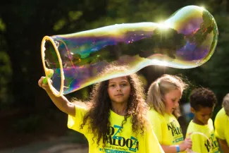 A young girl with long curly hair uses a wand to blow a giant bubble. There are trees and a group of other kids out of focus in the background.