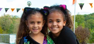 A close-up of two young girls smiling and standing close together. Both are wearing black cat ear headbands with cat ears. The girl on the left has long, straight hair and is wearing a black shirt with a bright, multi-colored animal print. The girl on the right has curly hair and is wearing a solid black shirt. In the background, there is a string of orange and black Halloween flags, and a landscape of trees under a cloudy sky.