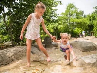A young girl holding hands with a toddler as they climb barefoot up a steep, rocky hill. They are surrounded by trees and large rocks.