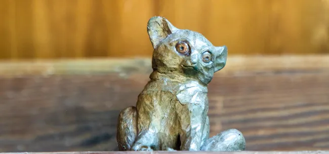 A small, bronze sculpture of a Bush Baby-a small, mousey looking primate with small, rounded ears and large, round eyes sitting in front of a wooden wall.