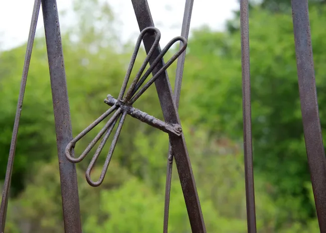 A closeup on a part of a metal art installation that showcases a small, metal dragonfly appearing mid flight in front of some tall cattail stalks. There is a green forest visible in the blurred background.