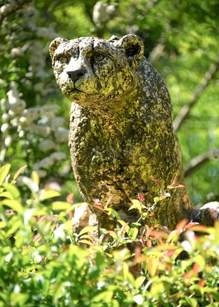 A bronze rendering of a large cat, possibly a Leopard as it sits, poised to strike on a rock surrounded by tall grasses under a clear blue sky.