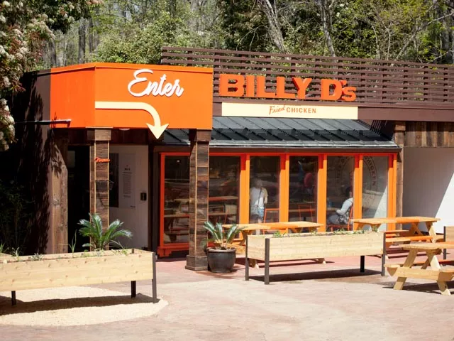 A rectangle shaped brown and orange building, flanked with flower beds and benches, with a large orange sign that reads "Billy D's Restaurant"