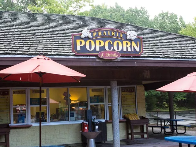 A small tan building with a brown roof that holds a sign that reads "Prairie Popcorn". There are red, outdoor umbrellas scattered around the building.